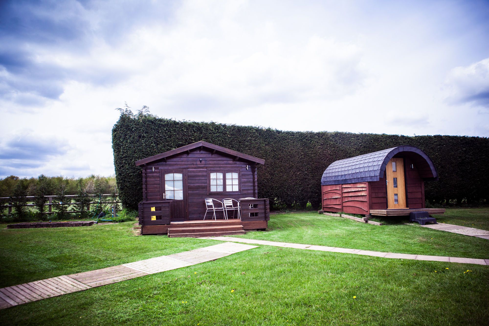Two wooden cabins with a paved walkway in between, surrounded by a well-maintained lawn and a hedge.