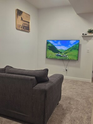 A living room with a flat-screen TV mounted on the wall, a dark-colored sofa, and a carpeted floor. - Charming 2-bedroom House Rental in Calgary - Welcome to Homely Harbour! (Calgary)