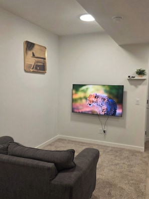 A living room with a flat-screen TV mounted on the wall, a gray sofa, and a small shelf with a plant. - Charming 2-bedroom House Rental in Calgary - Welcome to Homely Harbour! (Calgary)