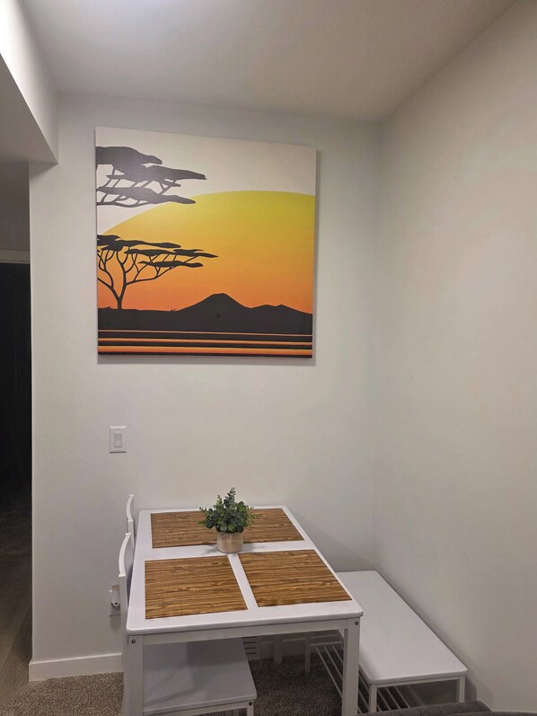 A room with a white table, a small bench, and a wall-mounted painting of a sunset. - Charming 2-bedroom House Rental in Calgary - Welcome to Homely Harbour! (Calgary)