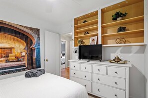 Ein Schlafzimmer mit einem Bett, einem Fernseher auf einer weißen Kommode, einem Wandregal mit Dekoration und einem großen Wandteppich.