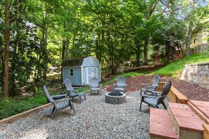 Eine Kiesterrasse mit Adirondack-Stühlen, einem kleinen Schuppen und einer Feuerstelle.
