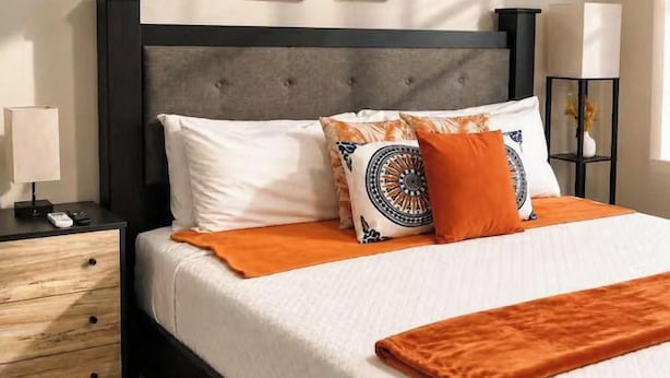 Una cama con cabecera gris, ropa de cama blanca y detalles naranja. Se observa un mesita de noche con una lámpara y un control remoto.