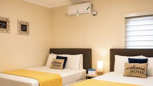 Habitación de hotel con dos camas, cada una con una almohada que dice 'bienvenido a casa', una mesita de noche con lámpara y un equipo de aire acondicionado en la pared.
