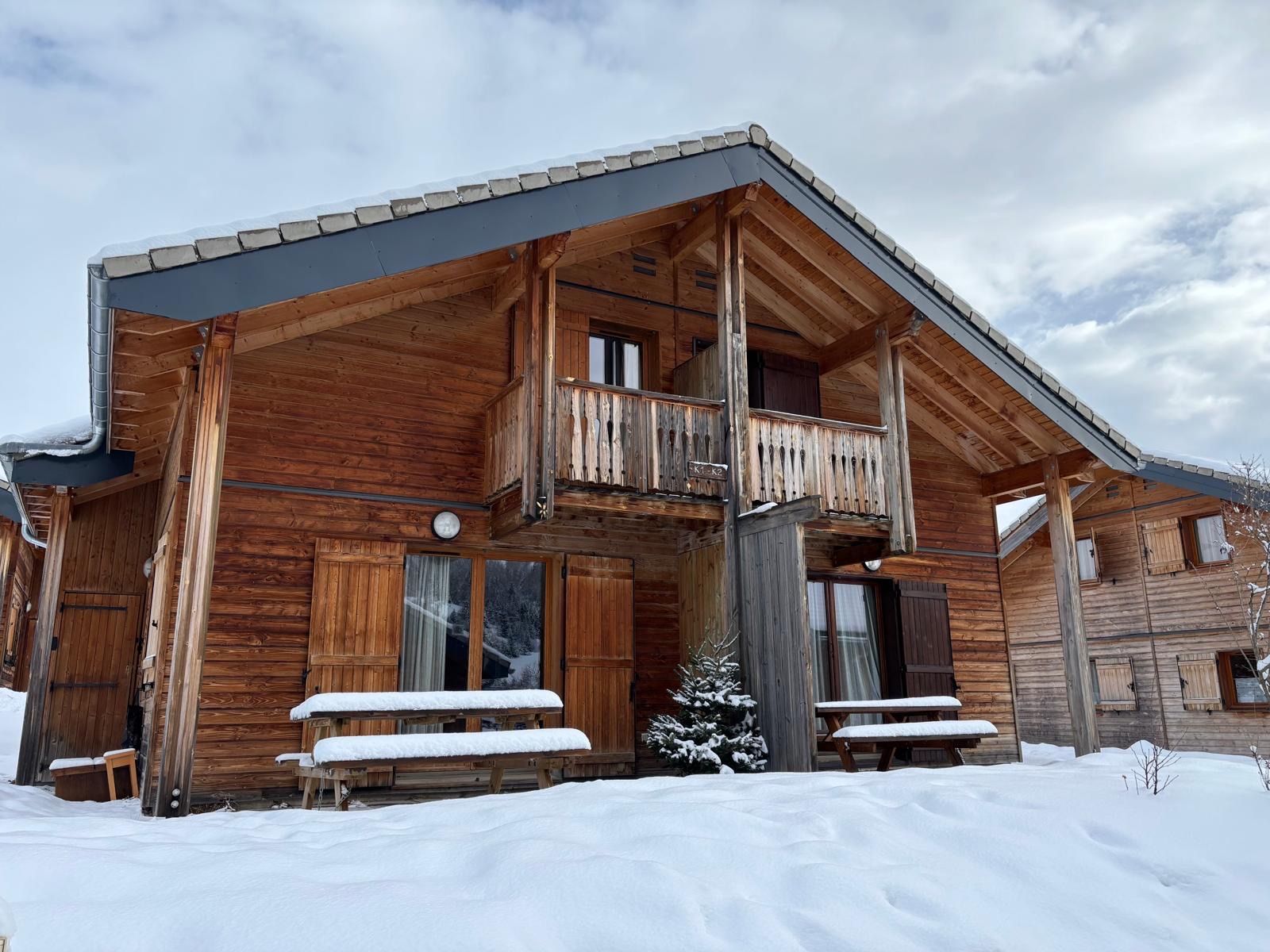 A wooden cabin with a balcony, snow-covered benches, and a small tree.