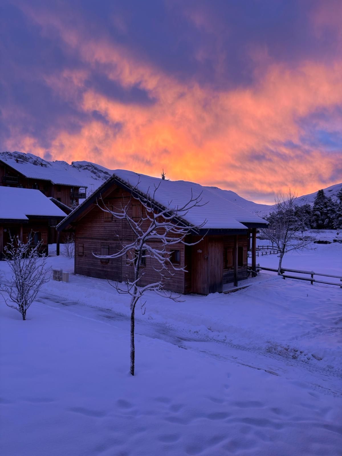 A wooden cabin with a snow-covered roof, surrounded by a snowy landscape and mountains in the background.