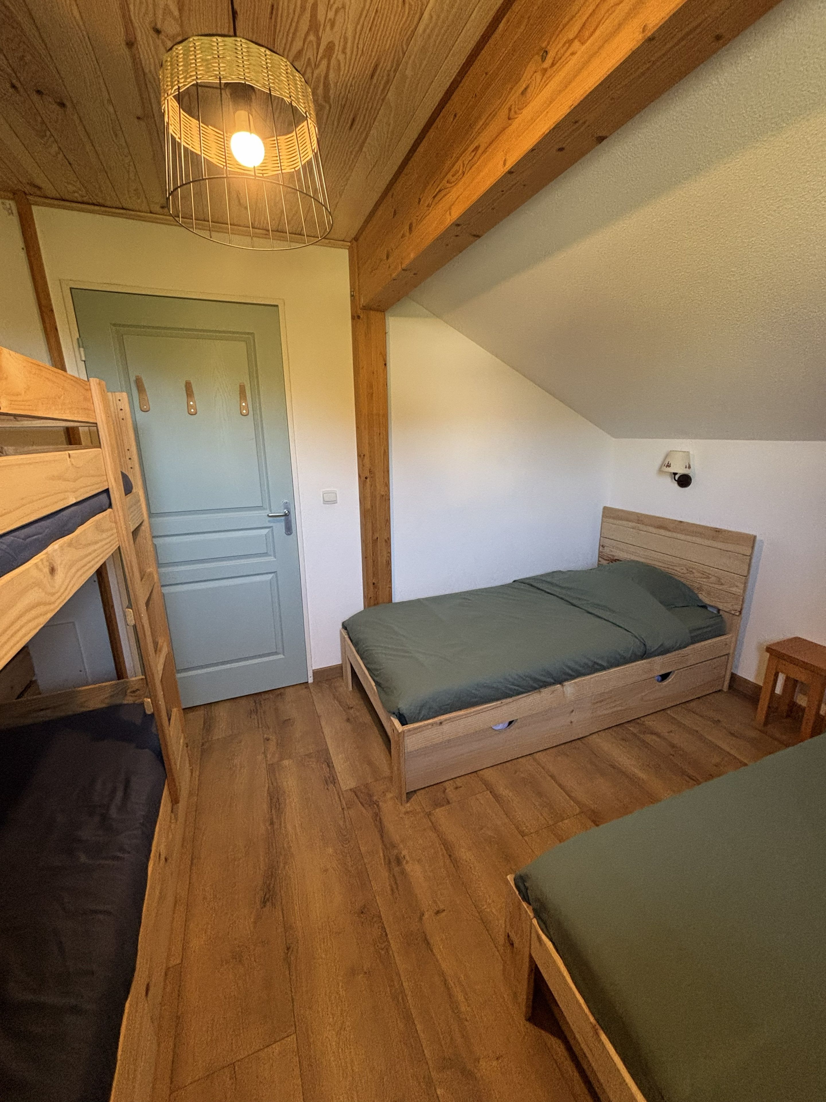 A room with a bunk bed, a single bed, a wooden ceiling, and a door.