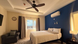 Una habitación de hotel con una cama, un ventilador de techo, una ventana con cortinas, un reloj y un equipo de aire acondicionado.