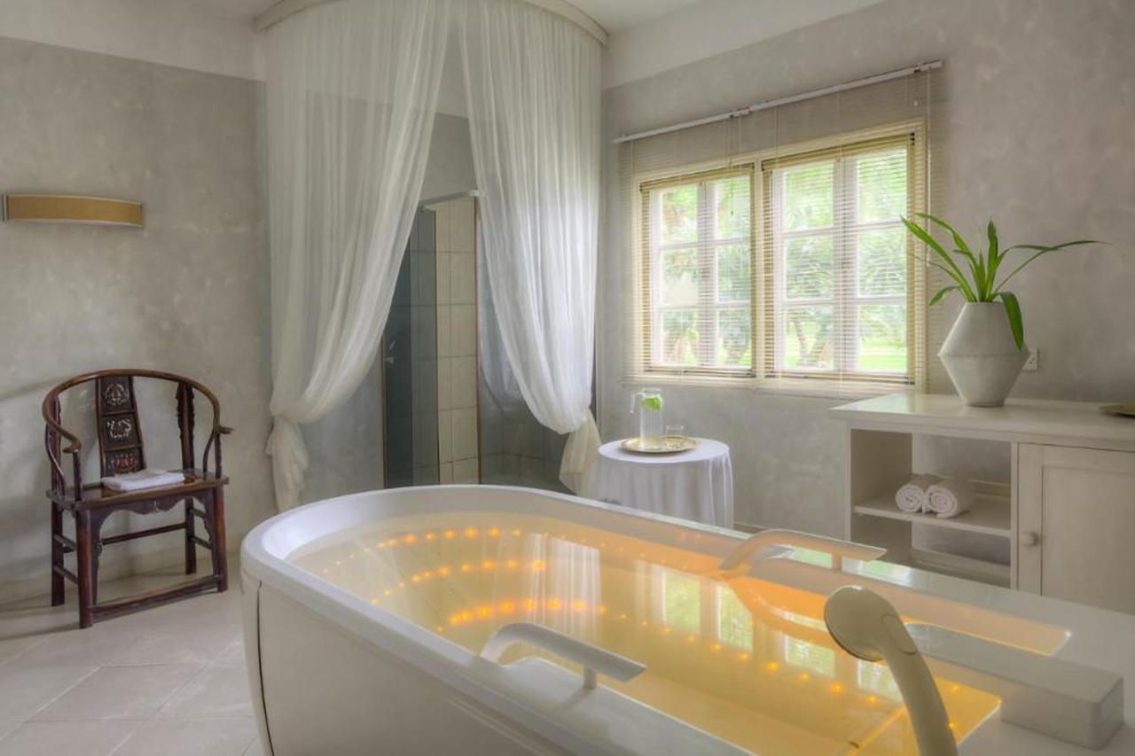 A bathroom with a bathtub, a wooden chair, a small table with a plant, and a window with blinds.