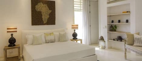 A bedroom with a large bed, bedside tables, a wall-mounted artwork, a bookshelf, and a window with curtains.