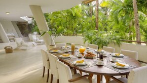 A dining area with a wooden table set for breakfast, surrounded by white chairs, with a view of lush greenery outside. - Lion In The Sun Hotel (Malindi)
