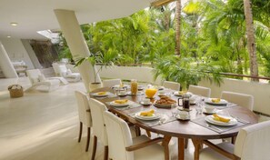 A dining area with a wooden table set for breakfast, surrounded by white chairs, with a view of lush greenery outside. - Lion In The Sun Hotel (Malindi)