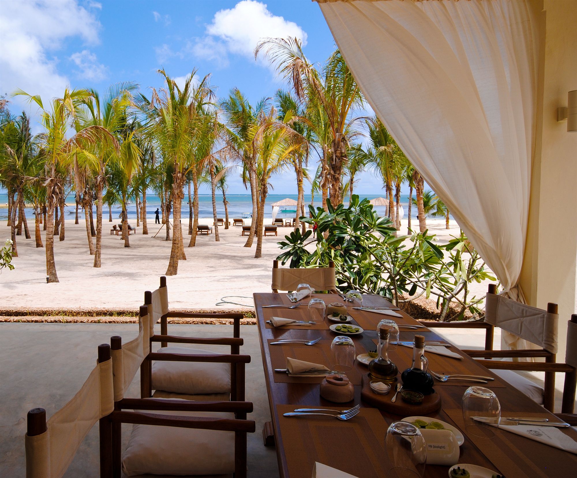 Un área de comedor frente a la playa, con una mesa para dos, con vista al océano y palmeras.