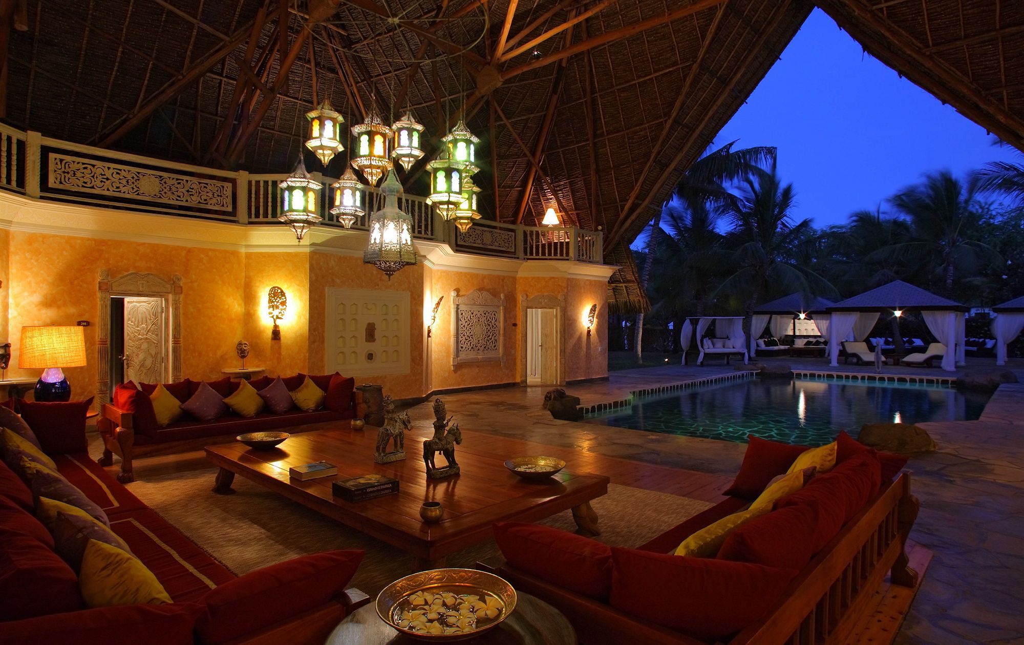 Elegante zona de lounge con piscina, techo de paja, iluminación ornamentada y decoración tropical.