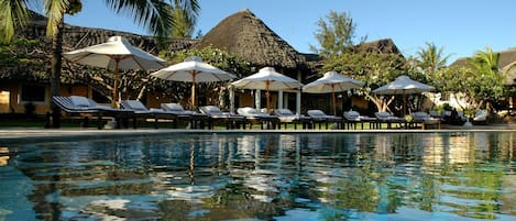 A pool area with lounge chairs and umbrellas, surrounded by palm trees and thatched-roof structures.