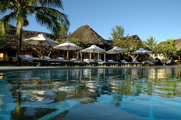A pool area with lounge chairs and umbrellas, surrounded by palm trees and thatched-roof structures. - Lion In The Sun Hotel (Malindi)