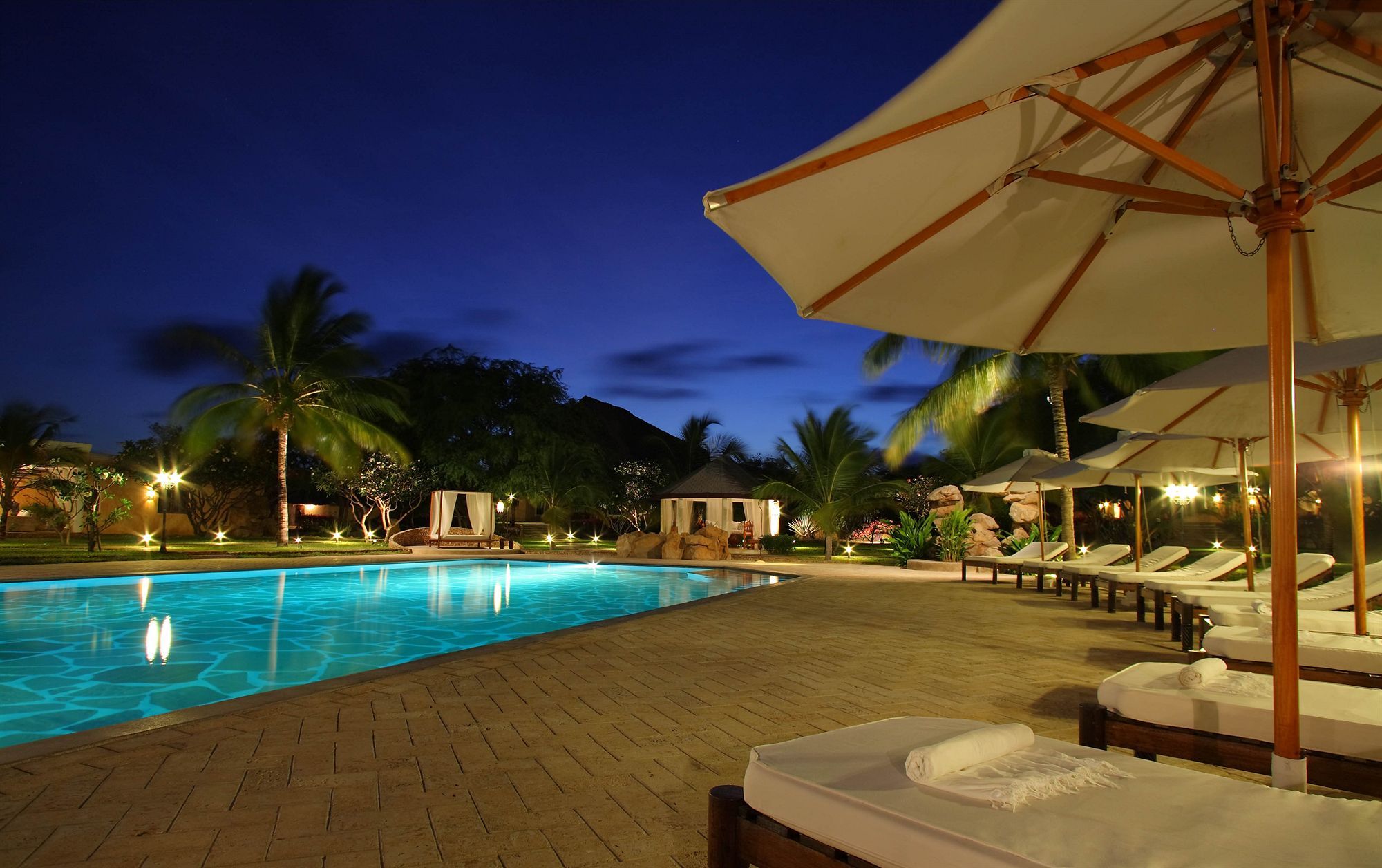 A swimming pool with lounge chairs and umbrellas, surrounded by palm trees and a night sky.