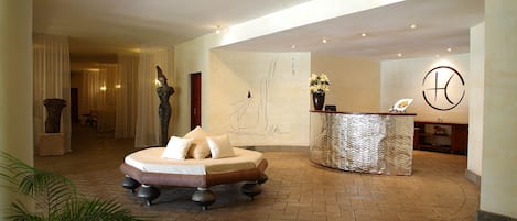 Vestíbulo de hotel con una mesa de recepción, una zona de recepción, un mural grande en la pared y un área de estar con sofá y cojines.