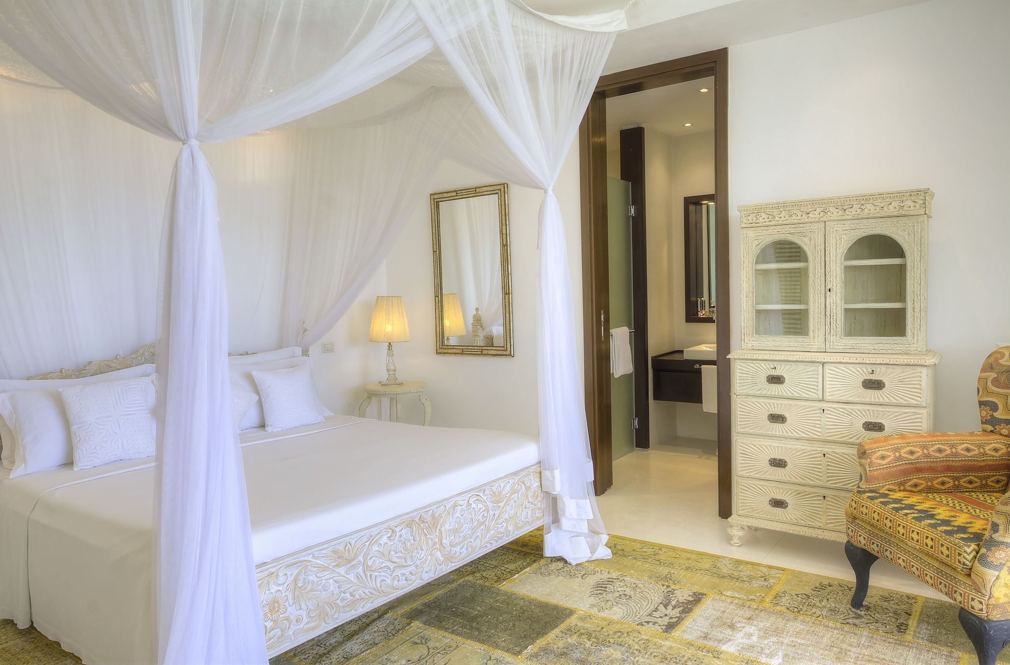 A bedroom with a four-poster bed, a bedside lamp, a mirror, and a cabinet with drawers.