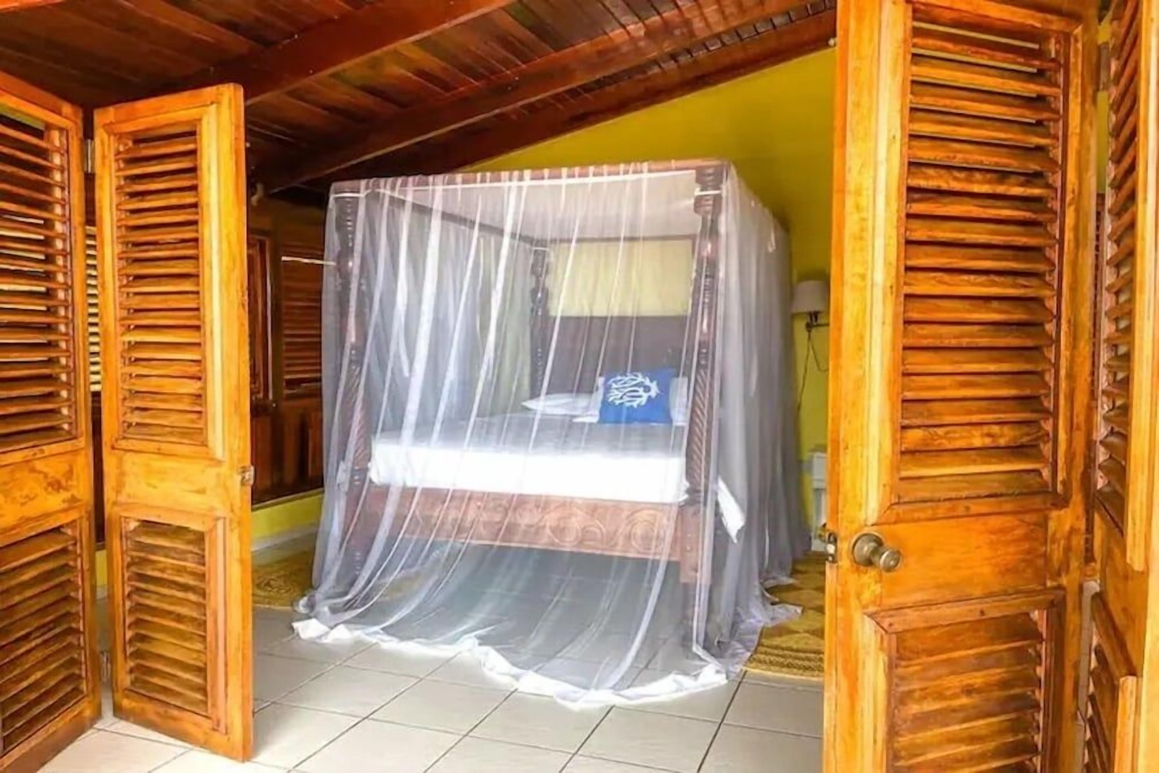 A room with a bed covered by a mosquito net, wooden shutters, and a tiled floor.
