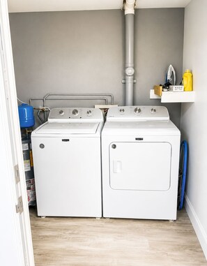 A laundry room with two front-loading washing machines, a water heater, and a shelf with laundry supplies. - Private Cottage | Walk to Beach, Tropical Setting (Delray Beach)