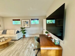 A modern living room with a large flat-screen TV, a sofa, a coffee table, and two potted plants. - Private Cottage | Walk to Beach, Tropical Setting (Delray Beach)