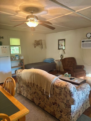 A living room with a ceiling fan, a couch, a chair, a table, a bed, a window, and a wall-mounted clock.