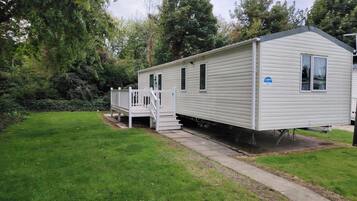 A mobile home with a front porch and a small deck, surrounded by a grassy yard and trees.