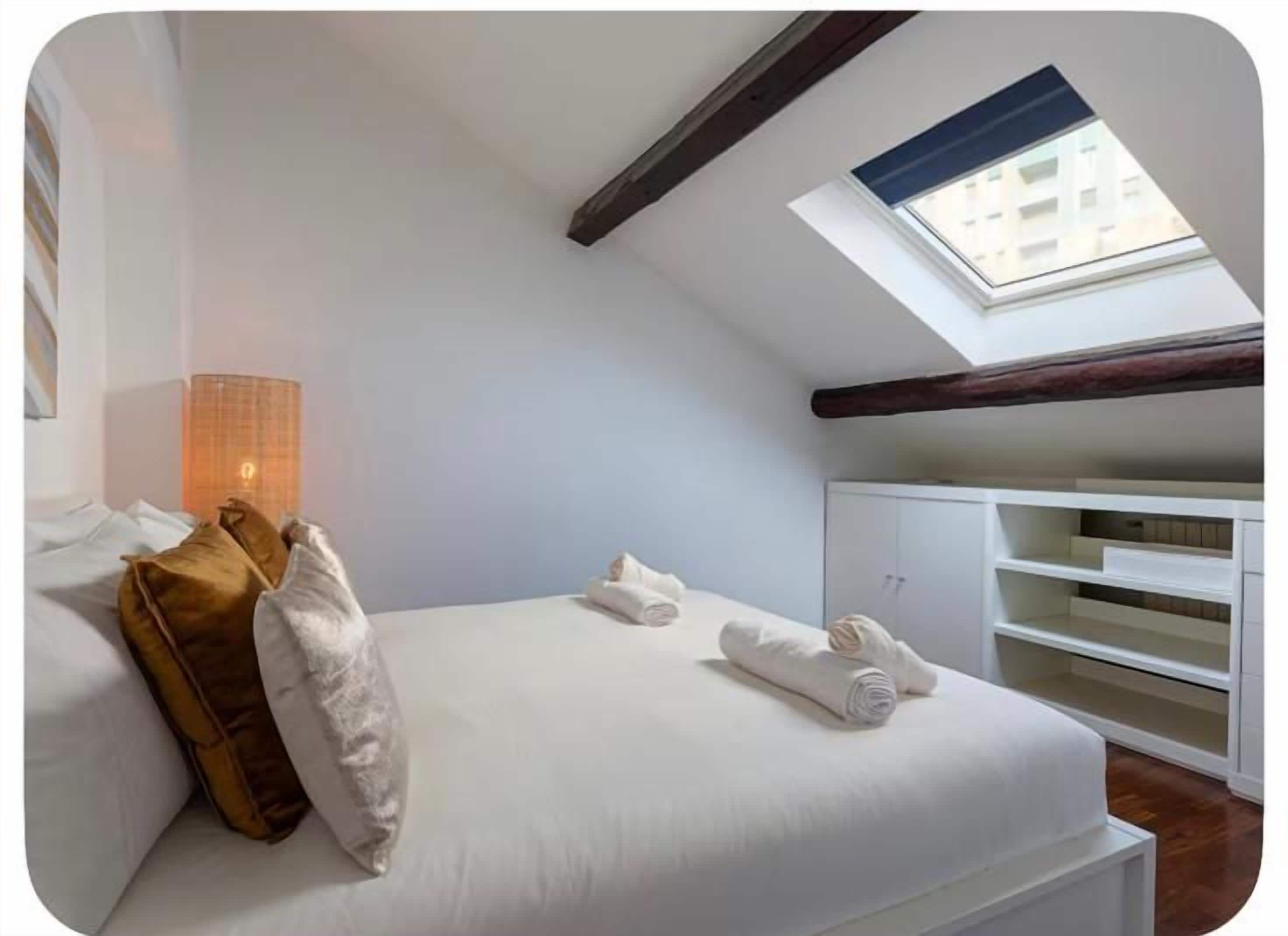 A bedroom with a skylight, a bed with pillows, a bedside lamp, and built-in shelving.