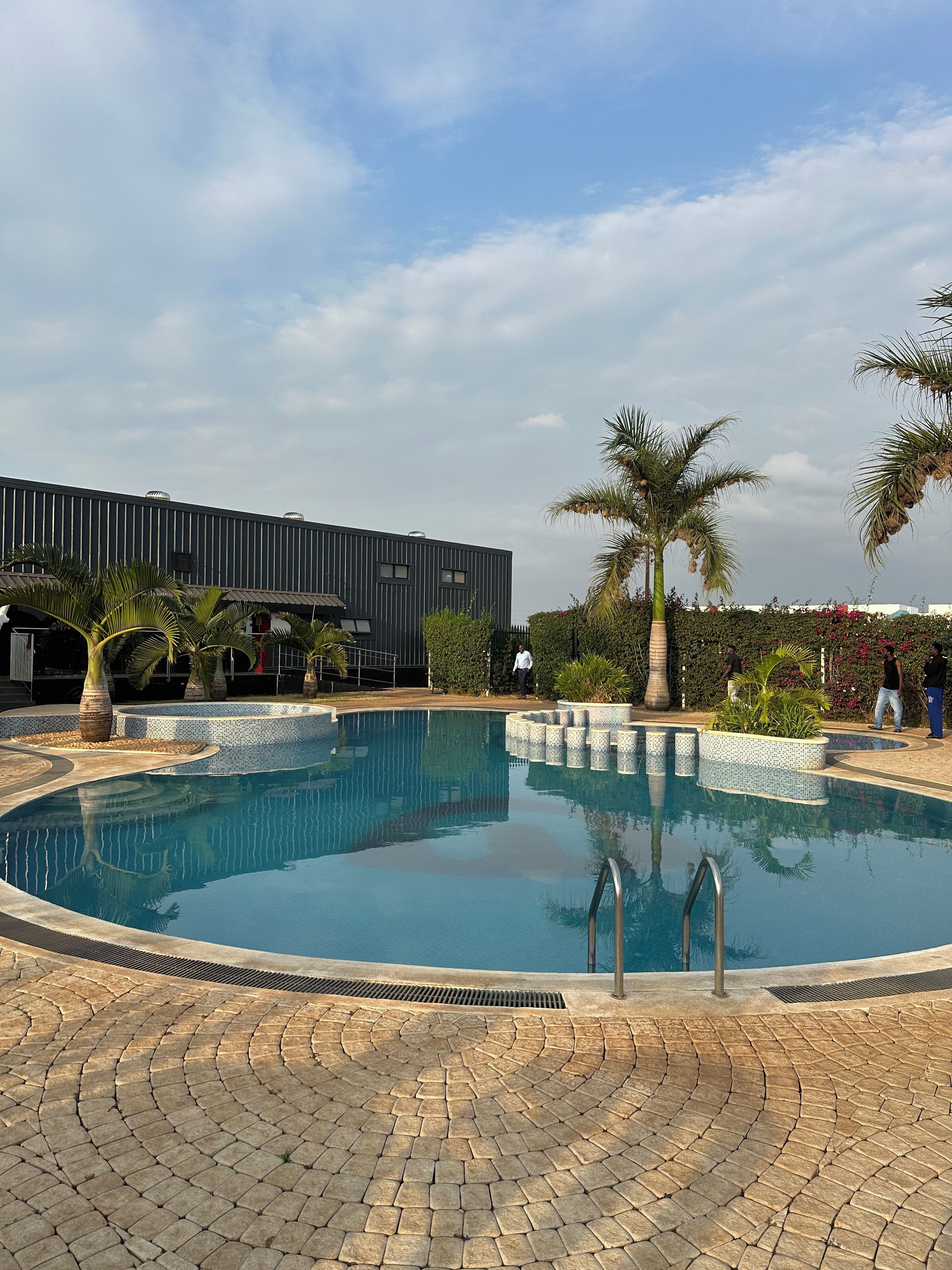 Une piscine avec une terrasse carrelée, des palmiers et un bâtiment moderne en arrière-plan.