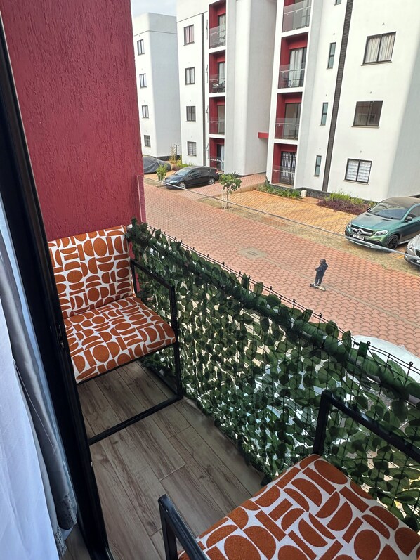 A balcony with a patterned cushion, a view of a residential area with parked cars, and a person walking.