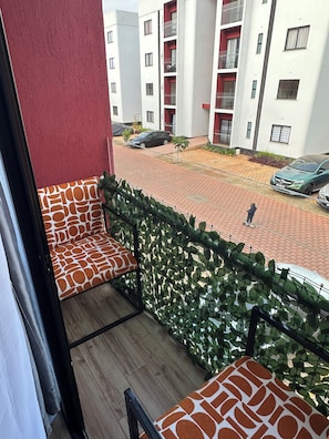 A balcony with a patterned cushion, a view of a residential area with parked cars, and a person walking.