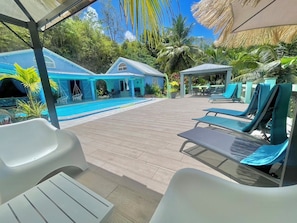 Un espace aménagé au bord de la piscine, avec des chaises longues, un coin salon abrité et un bâtiment doté d’un toit bleu.