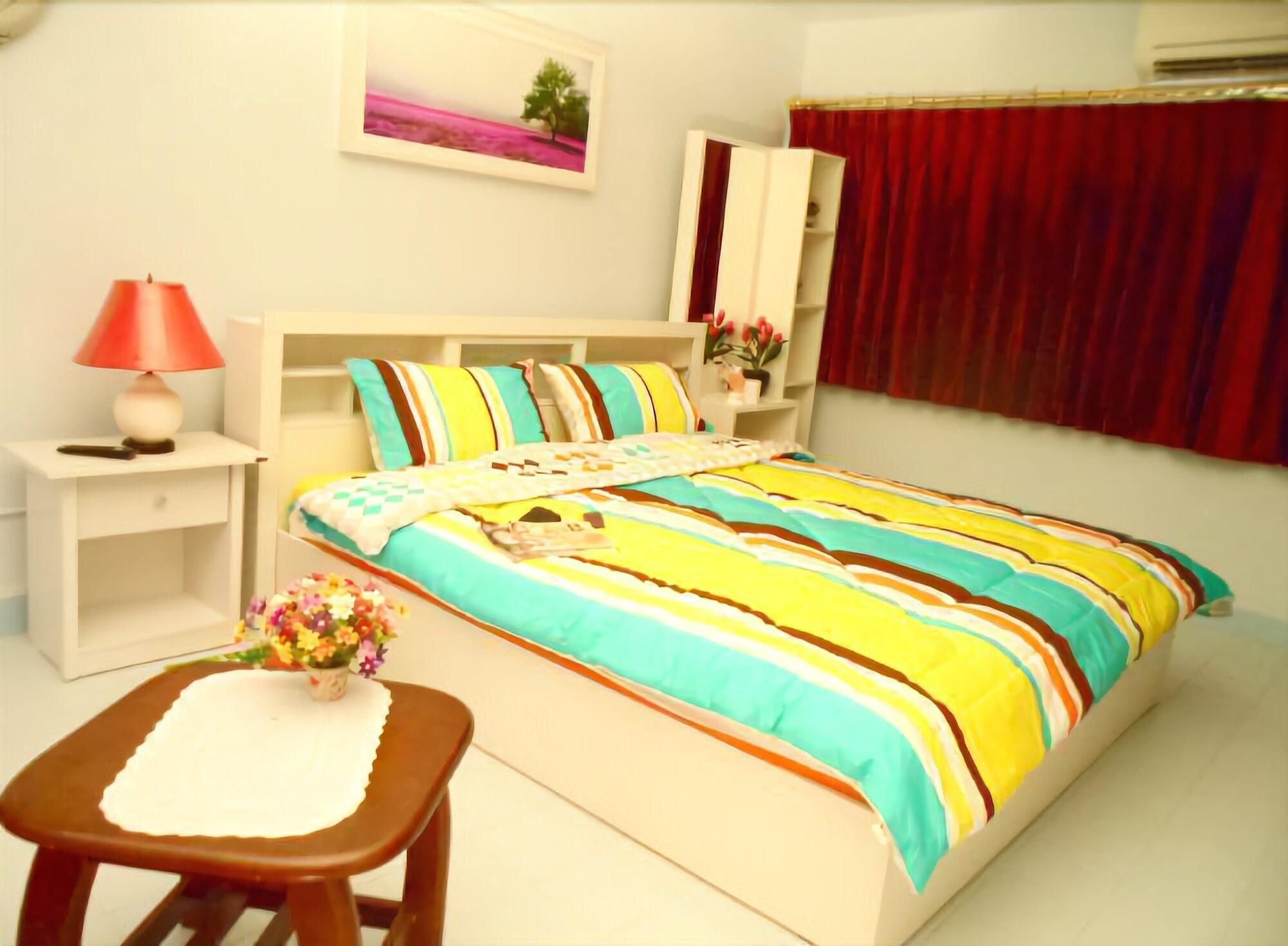 A bedroom with a bed, bedside tables, a nightstand, a chair, a lamp, a window with curtains, and a wall-mounted picture.