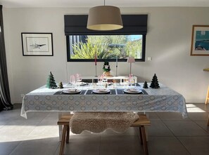 A dining area with a table set for four, a large window with a view, and a ceiling lamp. - Charming villa with a heated pool (Gujan-Mestras)