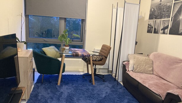 Una sala de estar con alfombra azul, un sillón verde, una silla marrón, un sofá rosa, una mesa de vidrio y una ventana con persianas.