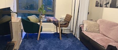 Un salon avec un tapis bleu, un fauteuil vert, une chaise marron, un canapé rose, une table en verre et une fenêtre avec des stores.