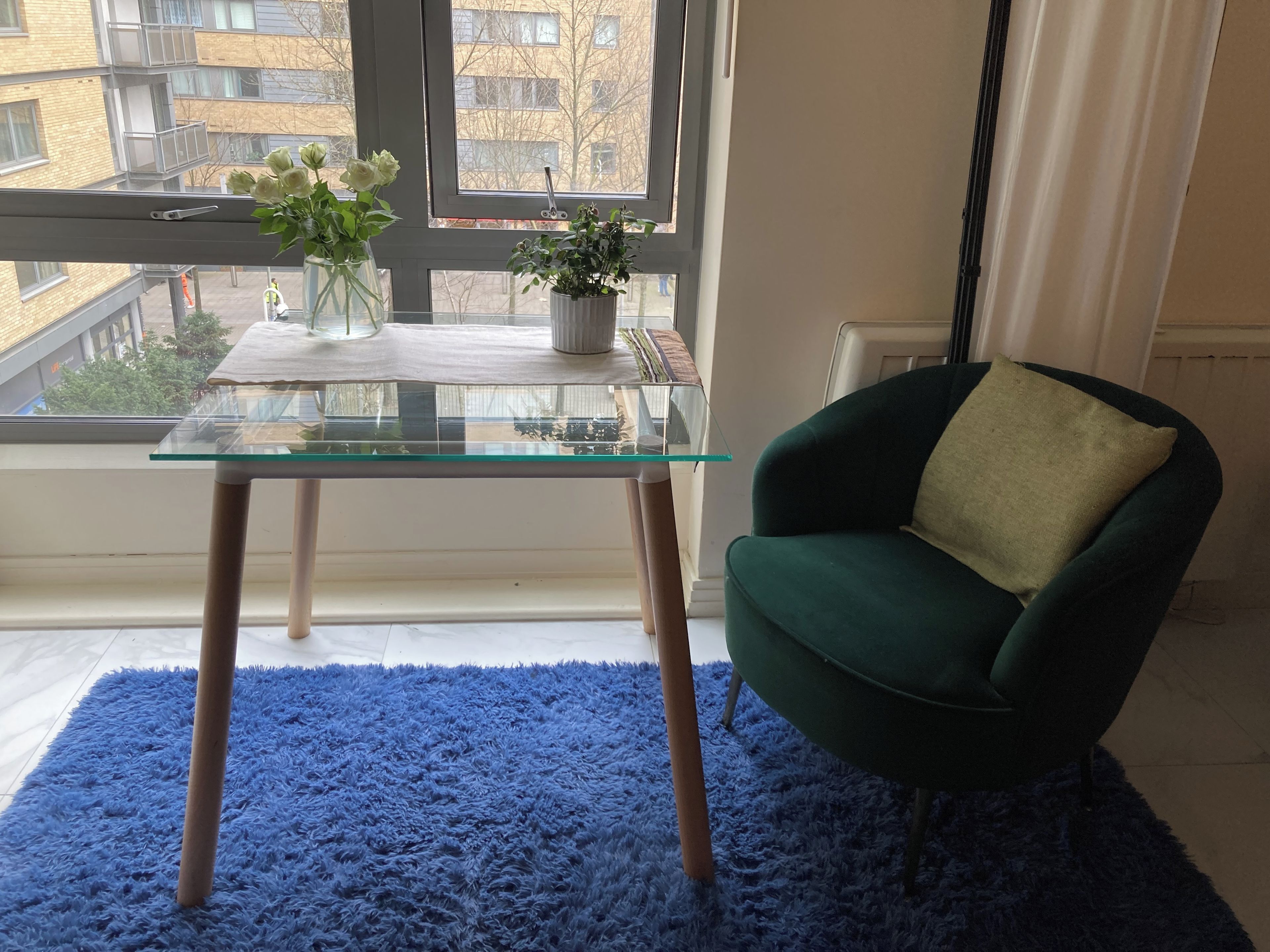 A room with a glass-top table, a green armchair, a blue rug, and a view of buildings through a window.