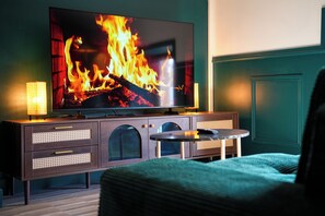 A room with a television displaying a fire, a cabinet with drawers, a round table, and a green wall.