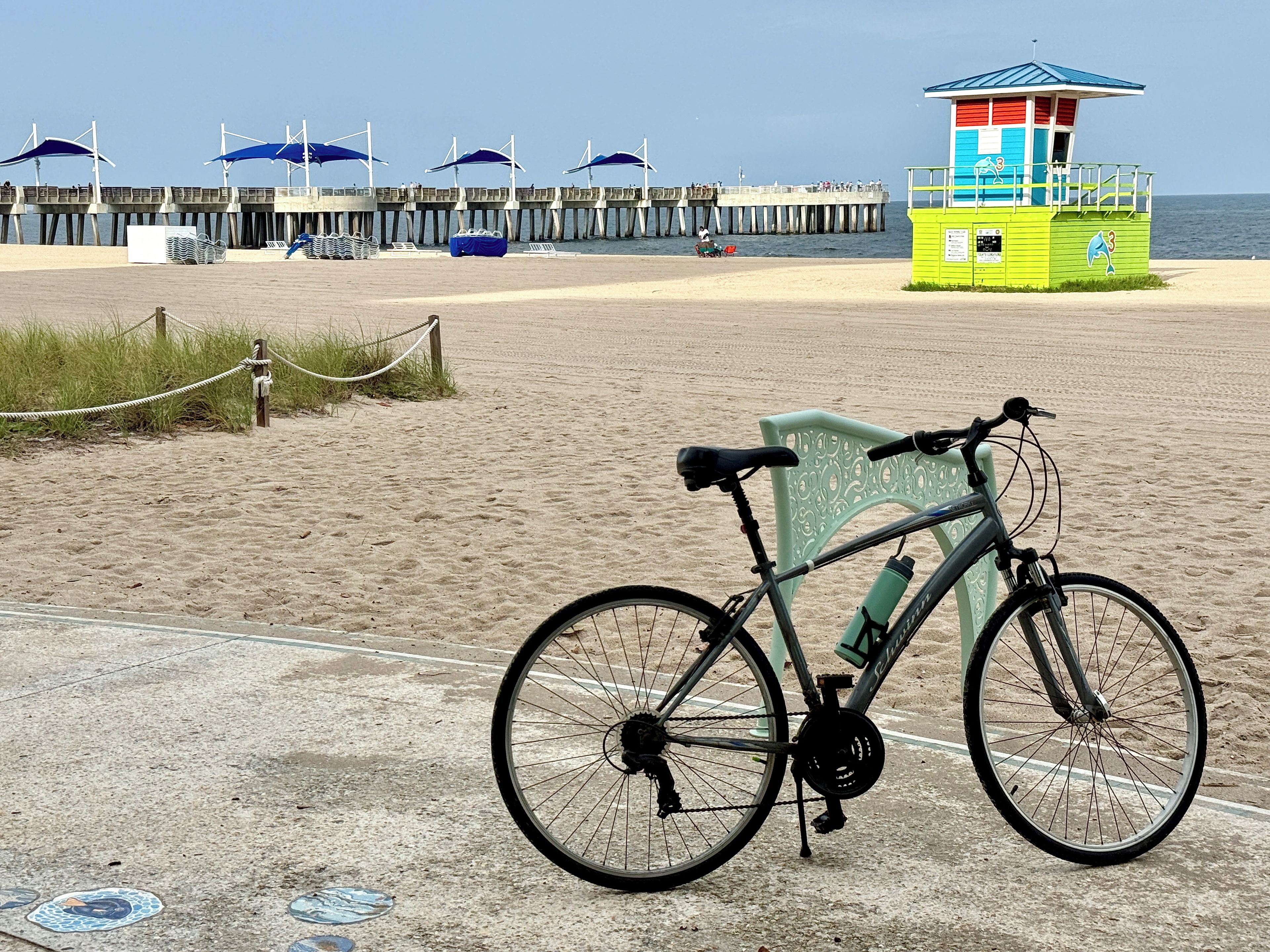 A beach with a bicycle parked on the sand, a lifeguard tower, and a pier with umbrellas.