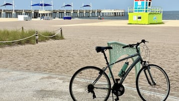 A beach with a bicycle parked on the sand, a lifeguard tower, and a pier with umbrellas.