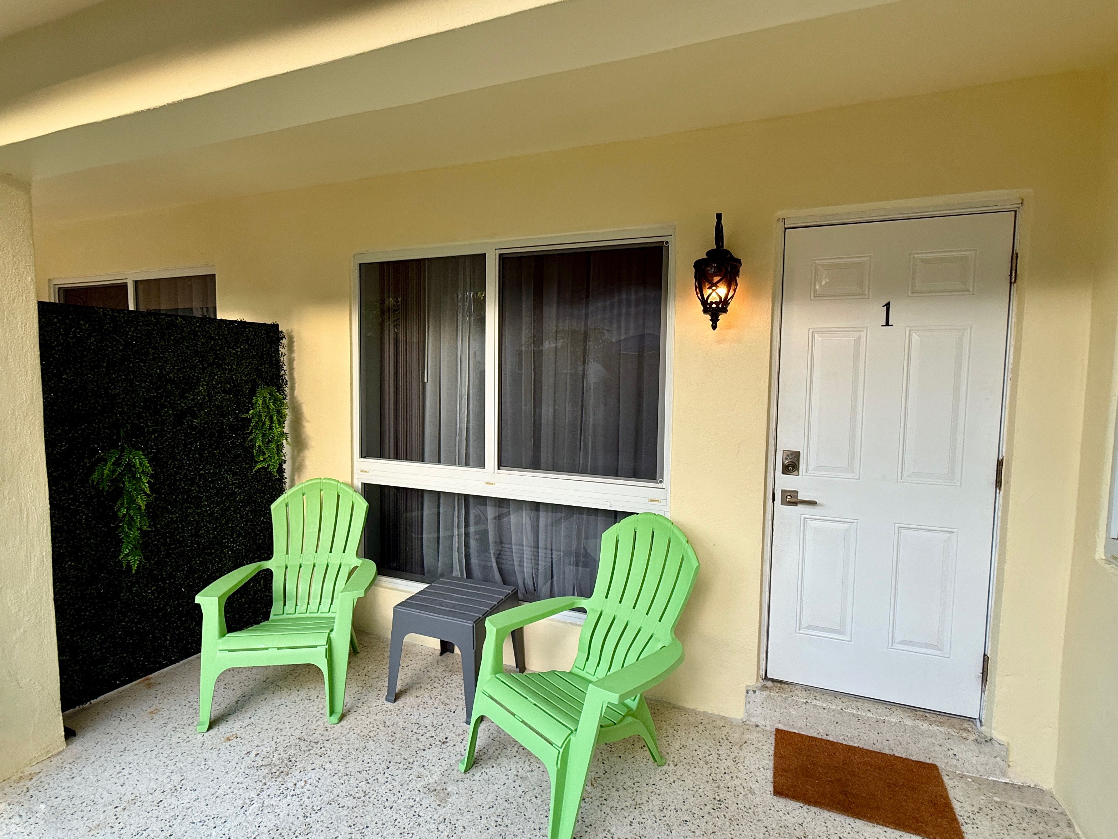 A front porch with two green chairs, a small table, a door marked with the number 1, and a wall-mounted light fixture.
