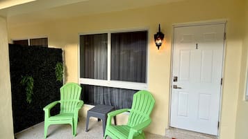 A front porch with two green chairs, a small table, a door marked with the number 1, and a wall-mounted light fixture.