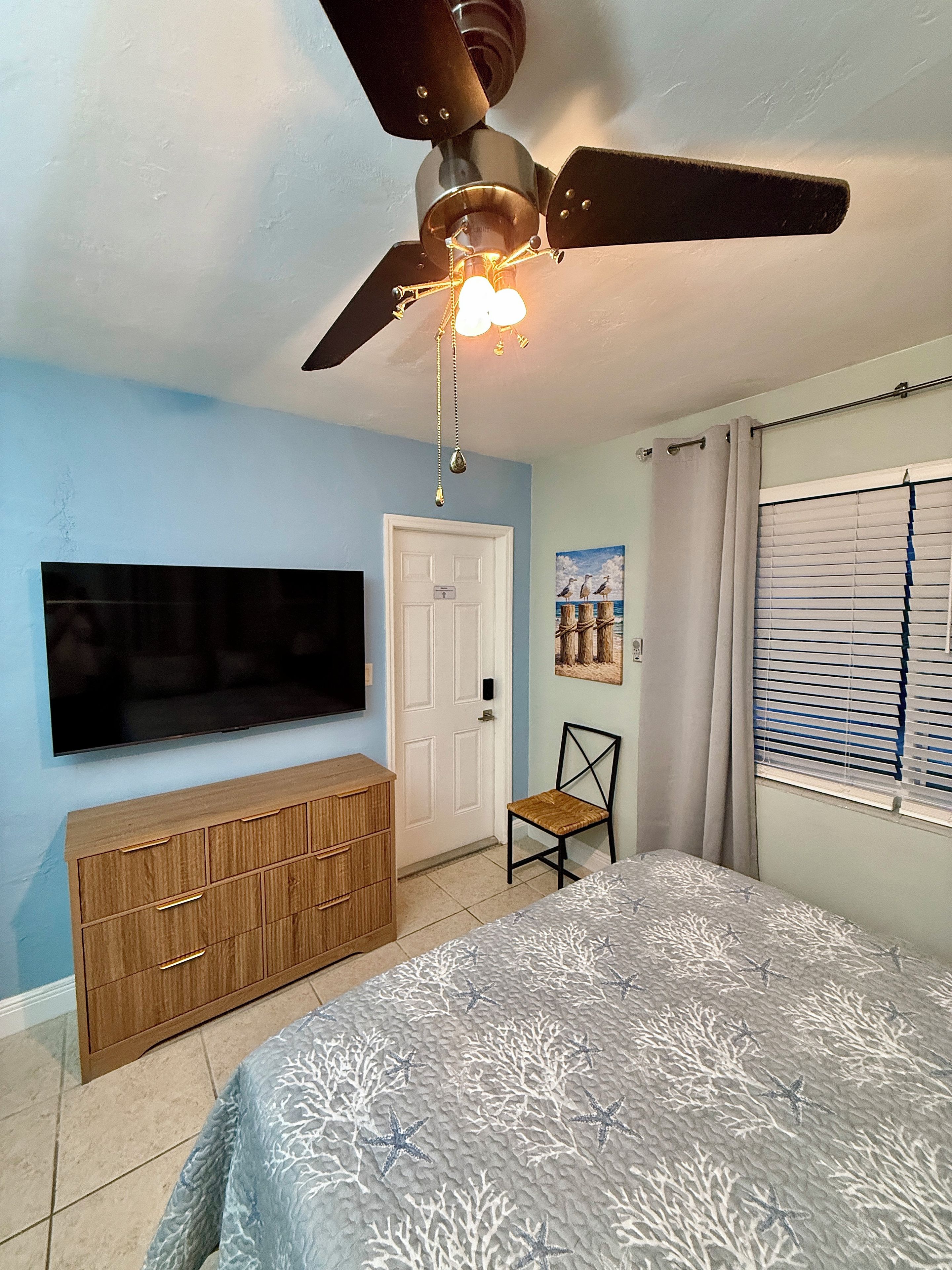 Un dormitorio con cama, televisor montado en la pared, ventilador de techo, cómoda de madera y una silla.