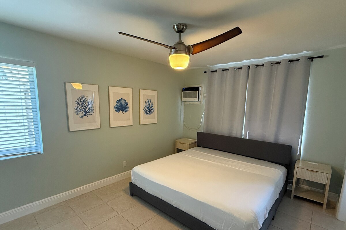 Ein Schlafzimmer mit einem Bett, einem Deckenventilator, einem Nachttisch, einer Wand-Klimaanlage und drei gerahmten Bildern.