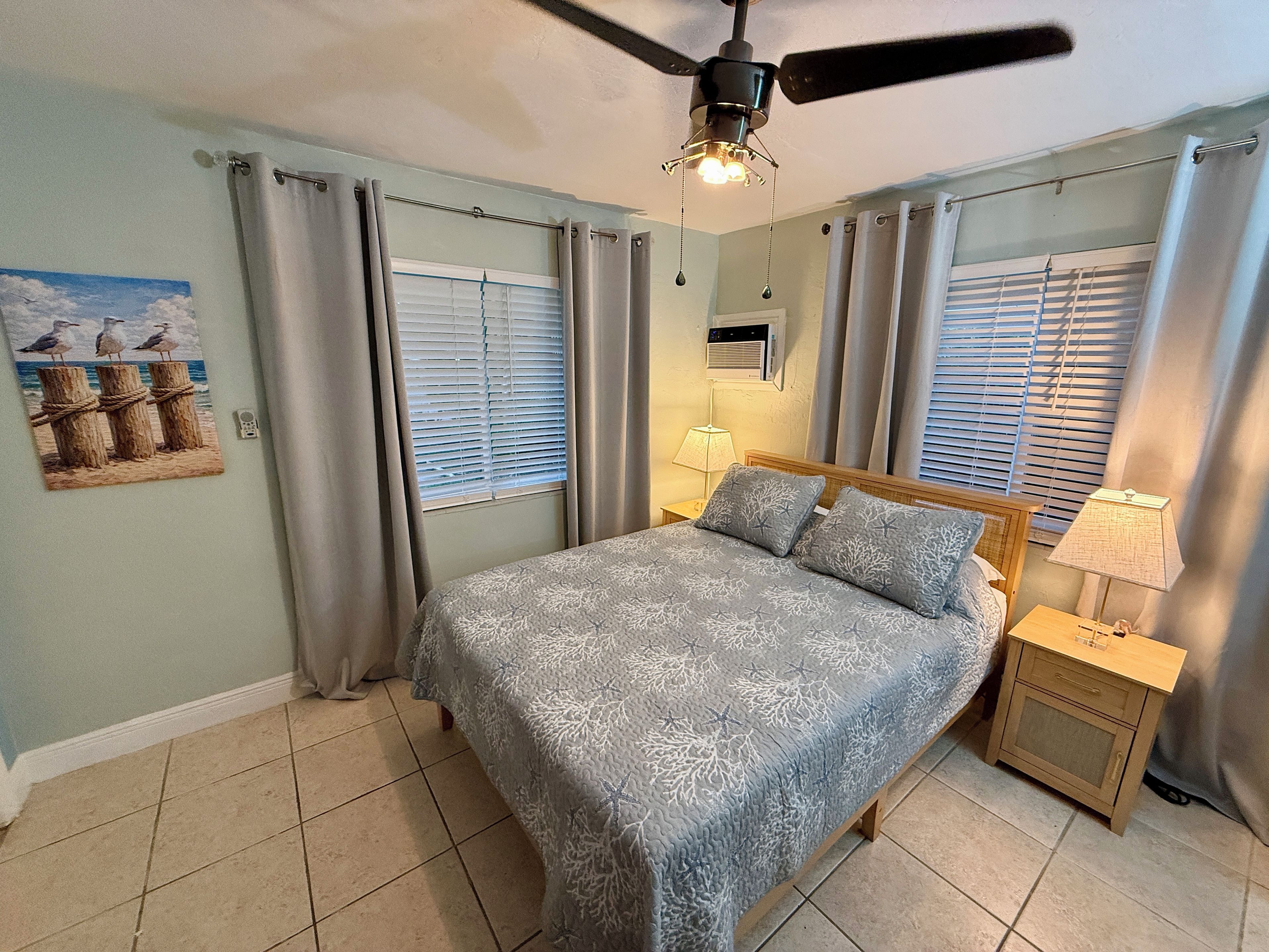 A bedroom with a bed, a ceiling fan, a nightstand with a lamp, and a wall-mounted air conditioner.