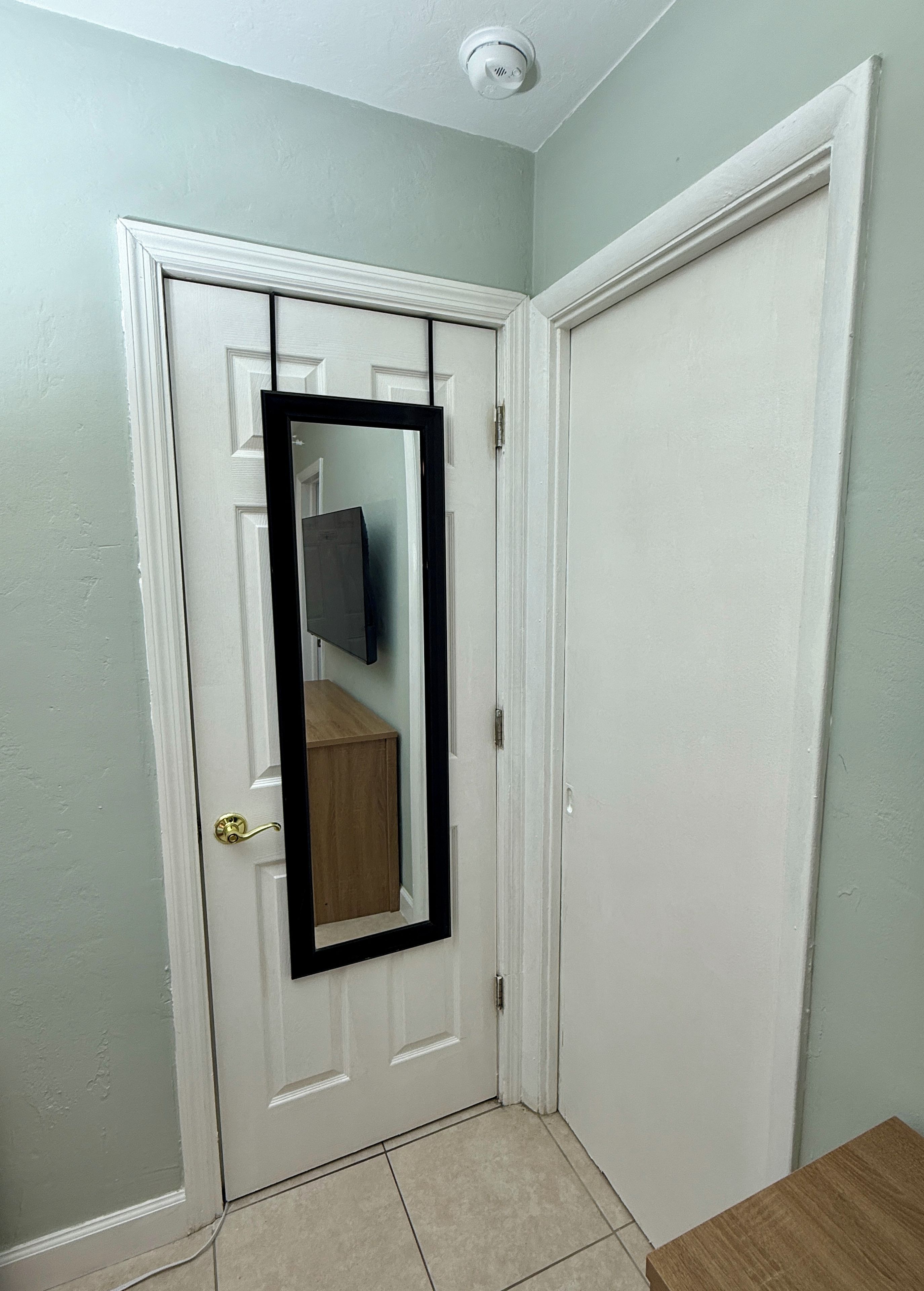 A white door with a black-framed mirror, a light green wall, and a wooden floor.