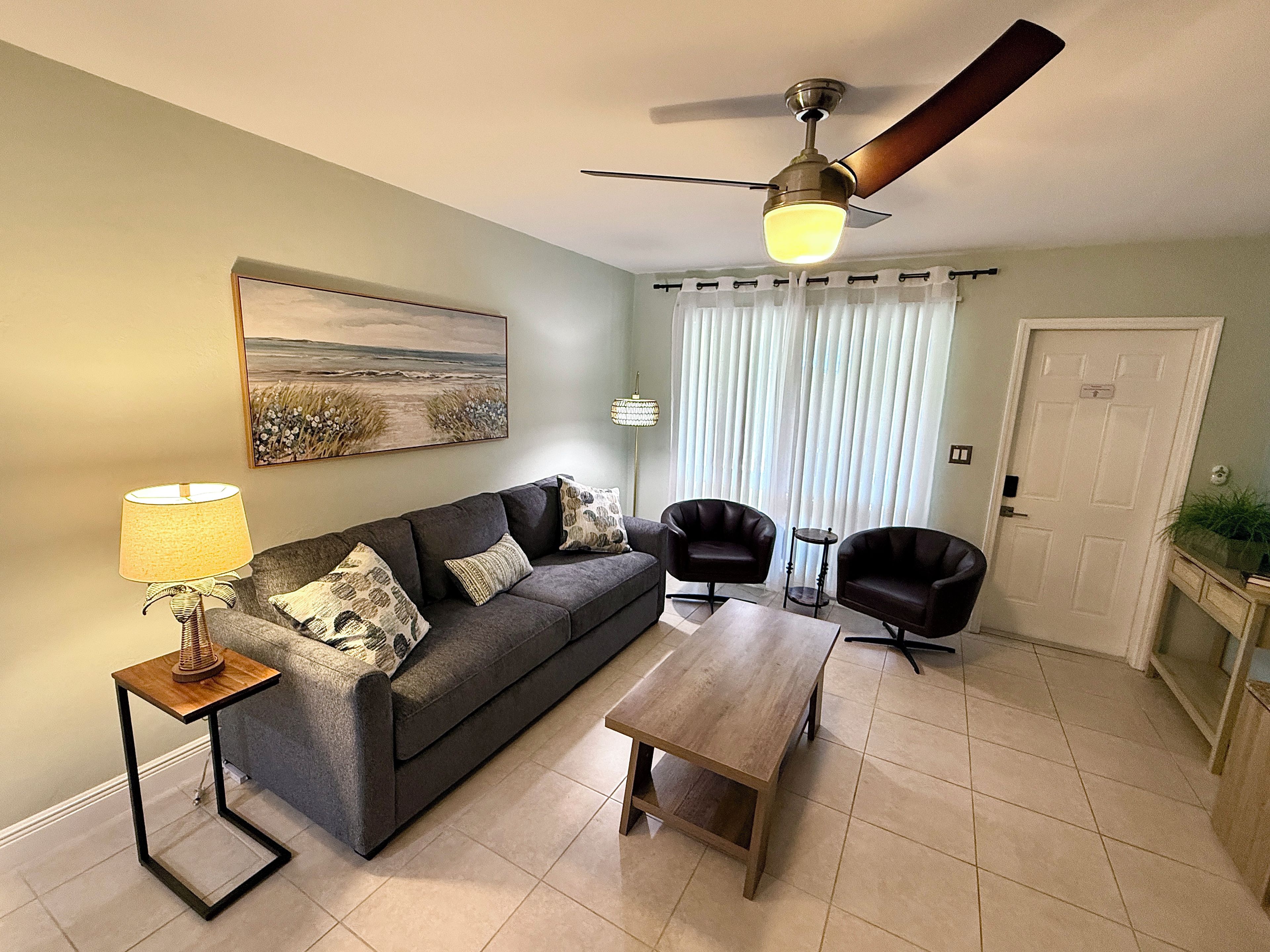 A living room with a sofa, two armchairs, a coffee table, a lamp, a ceiling fan, a painting, and a door.