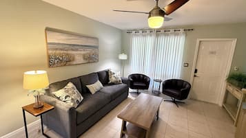 A living room with a sofa, two armchairs, a coffee table, a lamp, a ceiling fan, a painting, and a door.