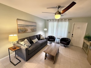 A living room with a sofa, two armchairs, a coffee table, a lamp, a ceiling fan, a painting, and a door. - Pompano Vibes Beach Apartments (Pompano Beach)